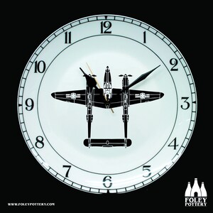May include: A white ceramic clock face with black numbers and hands. The clock face features a black silhouette of a P-38 Lightning aircraft. The clock is framed by a black ring with white numbers.