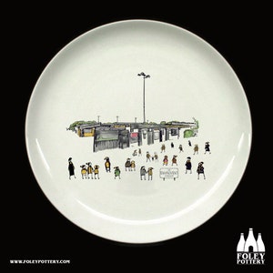 Könnte beinhalten: Ein weißer Keramikteller mit einer Schwarzweißillustration eines Stadions, auf dem Menschen zum Eingang gehen. Auf einem Schild steht der Text "The Environment Stadium".