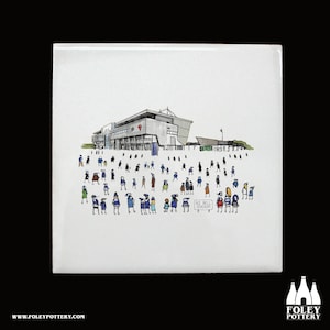 May include: A white ceramic tile featuring a black and white illustration of a stadium with a crowd of people in front of it. The stadium is a modern building with a large roof and a sign that reads "A.J. Bell Stadium".