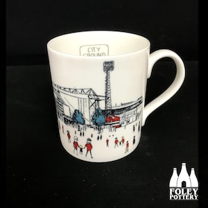 Puede incluir: Taza de cerámica blanca con una ilustración en blanco y negro de un estadio con el texto "City Ground" en la parte superior. La ilustración incluye figuras de palo de personas en rojo y azul.