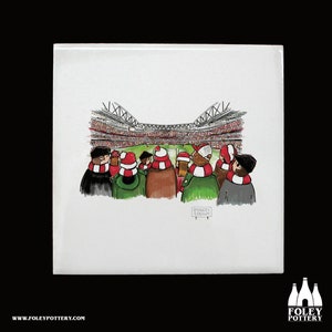 May include: A white ceramic coaster with a cartoon illustration of a crowd of people watching a soccer game at the Emirates Stadium. The people are wearing red and white scarves and hats.