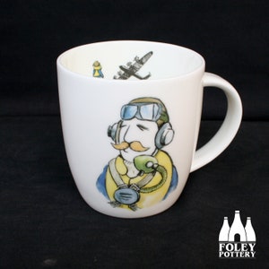 Könnte beinhalten: Weiße Keramiktasse mit einer Cartoon-Illustration eines Piloten mit Headset und Brille. Der Pilot hat einen Schnurrbart und schaut zur Seite. Die Tasse hat einen schwarzen Henkel und ein Flugzeug ist in der Tasse sichtbar.