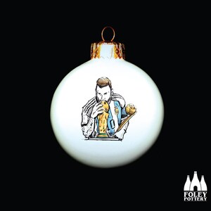 Può includere: Palloncino di Natale bianco con un'illustrazione disegnata a mano di un calciatore che tiene un trofeo. Il calciatore indossa una maglia blu e bianca e ha la barba.