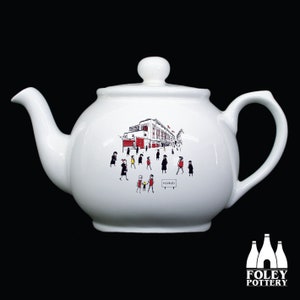 May include: White ceramic teapot featuring a black and white illustration of a stadium and people walking. The sign in the illustration reads "HIGHBURY".
