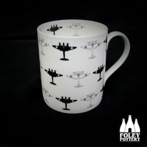 May include: A white ceramic mug with a black and white airplane pattern. The mug has a handle and is printed with the text "FOLEY POTTERY".