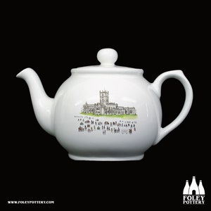 May include: White ceramic teapot with a hand-painted illustration of a church building and a crowd of people in front of it. The teapot has a rounded body, a spout, a handle, and a lid.