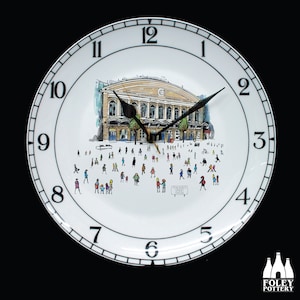 Puede incluir: Reloj de cerámica blanca con agujas y números negros. La esfera del reloj presenta una ilustración en blanco y negro de un edificio con personas caminando delante. El texto "Feachem Street Station" es visible en la esfera del reloj.