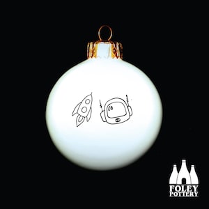 Könnte beinhalten: Weißer Weihnachtsbaumschmuck mit einer Schwarzweißillustration eines Astronauten und einer Rakete. Der Schmuck hat eine goldene Spitze.
