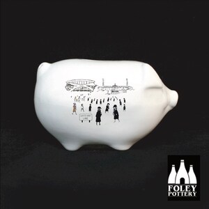 May include: White ceramic piggy bank with a black and white illustration of a stadium and people walking towards it. The illustration includes the text "White Hart Lane".