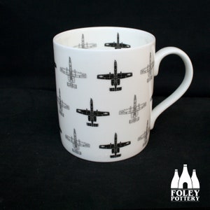 May include: White ceramic mug featuring a repeated pattern of black and white A-10 Thunderbolt II aircraft. The mug has a handle on the right side. The mug is made by Foley Pottery.