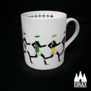 May include: White ceramic mug featuring a black and white illustration of runners wearing green, yellow and red jerseys. The mug has a black handle and is made by Foley Pottery.