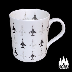 May include: White ceramic mug with a black and white pattern of fighter jets. The mug has a handle and the text "FOLEY POTTERY" is printed on the bottom.