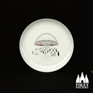 May include: White ceramic plate with a black and white illustration of a stadium with people walking towards it. The text "AFFINITY LEAGUE" is printed on a sign in the bottom left corner of the plate.
