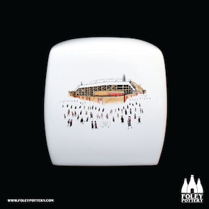 Può includere: Scatola in ceramica bianca con un'illustrazione dipinta a mano di uno stadio con persone che vi si dirigono. Lo stadio è marrone e rosso con un tetto bianco. Le persone indossano abiti neri e bianchi. Il testo "ROVER'S RETURN" è visibile sulla scatola.