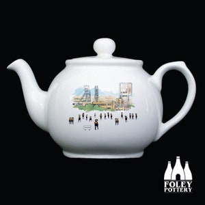May include: White ceramic teapot featuring a black and white illustration of a mine and miners. The teapot has a black handle and spout. The text "Florence" is visible on the teapot.