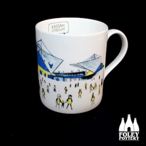 Op de afbeelding: Witte keramische mok met een handgetekend ontwerp van een voetbalstadion, met blauwe en gele kleuren. Het stadion is gelabeld "Kassam Stadium".