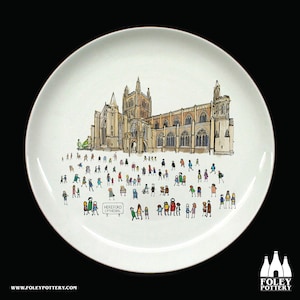 Puede incluir: Un plato de cerámica blanco con una ilustración dibujada a mano de la Catedral de Hereford y una multitud de personas frente a ella. El texto "HEREFORD CATHEDRAL" está impreso en un cartel frente a la multitud.