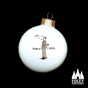Op de afbeelding: Witte kerstbal van keramiek met een zwart-witte illustratie van een man in een bruin pak en vest, die een pijp rookt, met vliegtuigen die erboven vliegen. De man staat voor een gebouw met een blauw dak. De kerstbal heeft een gouden top.