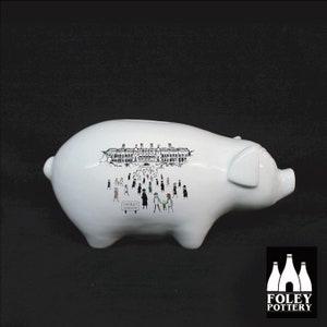 May include: White ceramic piggy bank with a black and white illustration of a building and people walking towards it. The building has the text "Gently Stadium" on a sign. The piggy bank is made by Foley Pottery.