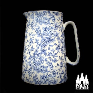 以下が含まれることがあります： 青い花柄の白いセラミックピッチャー。ピッチャーには大きなハンドルと注ぎ口が付いています。花柄のデザインはバラと葉が特徴です。Foley Potteryのロゴが右下に表示されています。