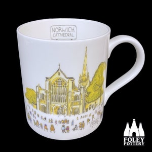 May include: White ceramic mug with a yellow and brown illustration of Norwich Cathedral and people walking in front of it. The text "Norwich Cathedral" is printed in gray on the top of the mug.