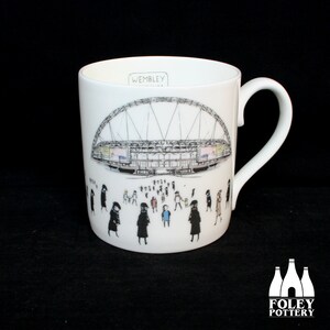 May include: White ceramic mug featuring a detailed illustration of Wembley Stadium. The design includes a crowd of people and the word "Wembley". The mug has a curved handle and a glossy finish. Foley Pottery logo is at the bottom.