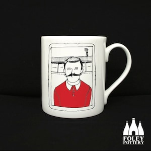 May include: White ceramic mug with a black and white illustration of a man with a mustache wearing a red jersey. The illustration is framed in a black border. The mug has a black handle.