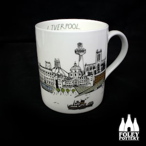May include: White ceramic mug featuring a black and green line drawing of the Liverpool skyline, including the Liver Building and the Radio City Tower. The word "Liverpool" is printed at the top. A Foley Pottery logo is at the bottom.