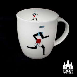May include: White ceramic mug featuring a black and white silhouette of a runner wearing a red, white and blue jersey. The mug has a rounded shape and a handle.