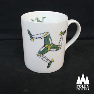 May include: White ceramic mug with a colorful design of the Isle of Man's three-legged symbol, known as the triskelion. The mug has a handle on the right side.