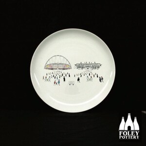 May include: White ceramic plate with a black and white illustration of a stadium and a crowd of people. The illustration is in a cartoon style. The plate is 10.5 inches in diameter.