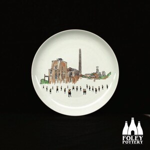 May include: A white ceramic plate with a hand-painted illustration of a factory and people in front of it. The factory has a tall chimney and is painted in shades of brown, gray, and green. The people are painted in black and yellow.