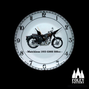 May include: White ceramic clock with black numbers and hands. The clock face features a black and white illustration of a Matchless 1955 G80S 500cc motorcycle. The text "Matchless 1955 G80S 500cc -" is printed below the motorcycle.