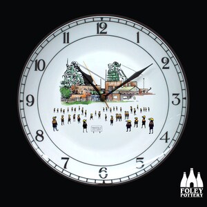 May include: A white ceramic clock with a black clock hand and black numbers. The clock face features a black and white illustration of a coal mine with the text "Mansfield Colliery". There are small black and yellow figures standing in front of the mine.