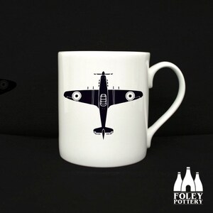Può includere: Una tazza in ceramica bianca con una silhouette nera di un aereo vintage stampata sul davanti. La tazza ha un manico sul lato destro.