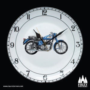 Puede incluir: Un plato de reloj de cerámica blanca con marcadores de hora romanos negros y manecillas negras. Una motocicleta azul está en el centro del plato. La esfera del reloj tiene un borde negro con un fondo blanco. La motocicleta es de estilo clásico con un tubo de escape cromado y un asiento negro. El reloj está hecho por Foley Pottery.