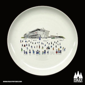 May include: A white ceramic plate featuring a black and white illustration of a stadium with a crowd of people in front of it. The text "A.J. Bell Stadium" is on a sign in the bottom right corner of the plate.