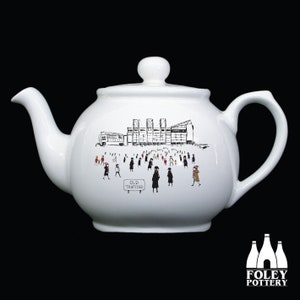 May include: White ceramic teapot with a black and white illustration of a stadium and people walking towards it. The text "Old Trafford" is on a sign in the image.