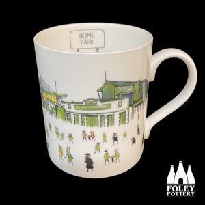Puede incluir: Taza de cerámica blanca con una ilustración verde y amarilla de un estadio con las palabras "Home Park" en un cartel encima. La taza tiene un asa blanca y un logotipo negro en la parte inferior que dice "Foley Pottery".