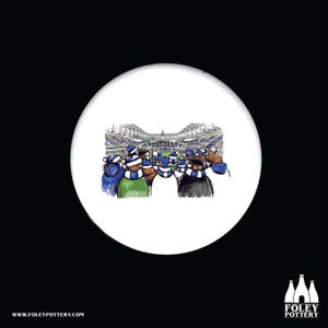 May include: A white ceramic round coaster with a black and white illustration of a crowd of people wearing blue and white hats watching a game in a stadium.