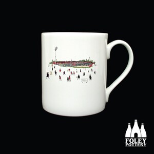 May include: White ceramic mug with a black and white illustration of a stadium with people in the foreground. The stadium is red and white. The text "Peninsula Stadium" is visible on the mug.