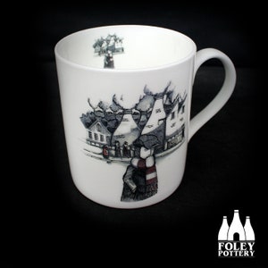 May include: White ceramic mug with a black and white illustration of a town with chimneys and a person wearing a red and white scarf. The mug has a black handle.