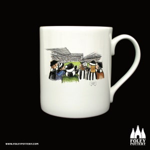 May include: White ceramic mug with a cartoon illustration of people watching a soccer game at St. James' Park. The mug has a black handle and the words "St. James' Park" are printed on the bottom of the mug.