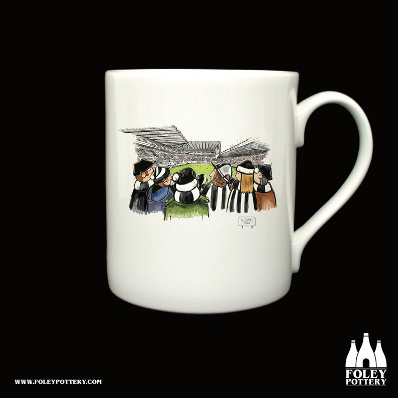 May include: White ceramic mug with a cartoon illustration of people watching a soccer game at St. James' Park. The mug has a black handle and the words "St. James' Park" are printed on the bottom of the mug.
