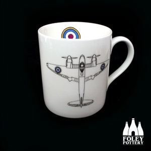 May include: White ceramic mug with a black and white illustration of a vintage airplane with a red, white, and blue roundel on each wing. The mug has a blue, white, and yellow roundel on the inside rim.