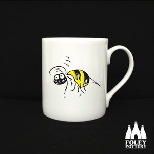 May include: White ceramic mug featuring a cartoon bee with yellow and black stripes. The bee is drawn with black outlines and is buzzing. The mug has a curved handle and a glossy finish. The Foley Pottery logo is at the bottom.