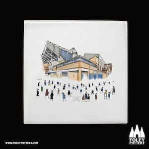 Puede incluir: Un azulejo de cerámica blanco con una ilustración dibujada a mano de un estadio de fútbol con una multitud de personas delante. El estadio está dibujado en negro, marrón y azul. Las personas están dibujadas en negro, azul y blanco. El azulejo tiene las palabras "Filbert Street" escritas en él.