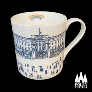 Puede incluir: Taza de cerámica blanca con una ilustración en blanco y negro del Palacio de Buckingham y un arcoíris en la parte superior. La taza tiene un asa negra y un fondo blanco. La ilustración incluye personas caminando frente al palacio.