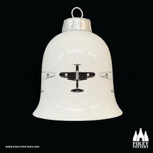 May include: White ceramic Christmas bauble shaped like a bell with a black silhouette of an aeroplane on the front. The bauble has a silver top and a silver band around the middle.