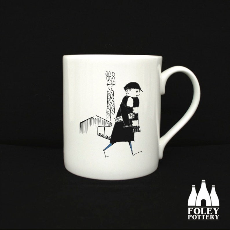 May include: White ceramic mug with a black and white illustration of a person wearing a scarf and walking past a building with a tall tower. The mug has a black handle and the words "FOLEY POTTERY" are printed on the bottom.